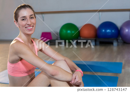 Woman sitting cross?legged on blue mat in yoga studio, smiling with balls and barre, copy space 128743147