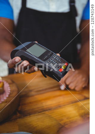 African American woman holding out card terminal at wooden cafe counter, with pink cake stand African American woman holding out card terminal at wooden cafe counter, with pink cake stand 128743149