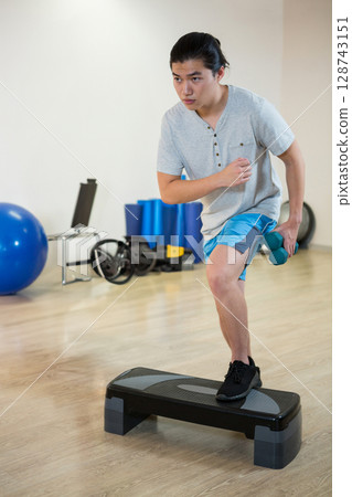 Asian young adult woman stepping onto aerobic step in indoor fitness studio, holding teal dumbbells 128743151