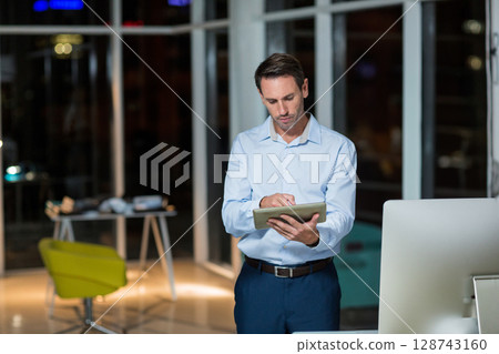 Businessman interacting with tablet at modern office at night, with desktop monitor and green chair 128743160