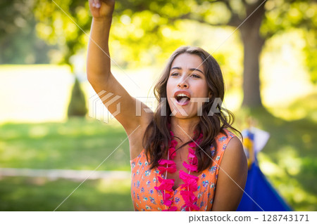 Woman raising arm and cheering in sunlit park, with pink flower lei at outdoor gathering Woman raising arm and cheering in sunlit park, with pink flower lei at outdoor gathering 128743171