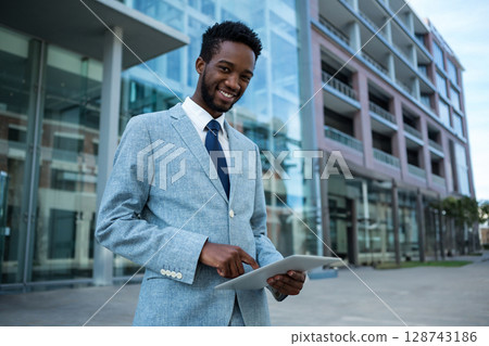 Young African American man standing outside office complex, smiling and tapping tablet computer Young African American man standing outside office complex, smiling and tapping tablet computer 128743186