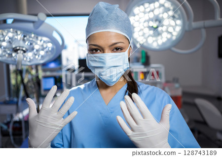 Female surgeon fitting gloves and scrub cap in operating room, with overhead lights and monitors 128743189