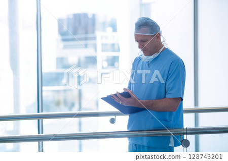 Man standing in hospital corridor reviewing clipboard near railing with surgical mask, copy space 128743201