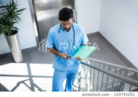 Asian man descending hospital stairwell using smartphone and carrying file folders with stethoscope 128743202
