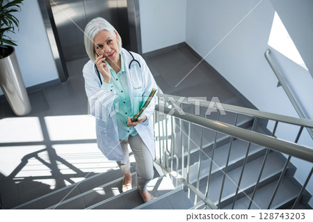 Senior woman doctor climbing stairs in hospital, talking on smartphone and holding patient files 128743203