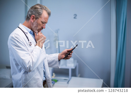 Middle-aged male doctor examining tablet in exam room, wearing stethoscope, IV stand, copy space 128743218