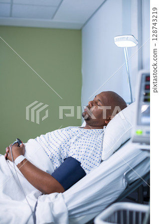 African American man lying in hospital room, with blood pressure cuff and bedside lamp 128743219