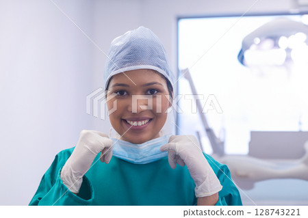 Female medical worker pulling down face mask in clinic room, wearing green scrubs and gloves 128743221