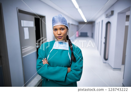 Female surgeon standing with arms crossed in hospital corridor, wearing green scrubs cap mask 128743222