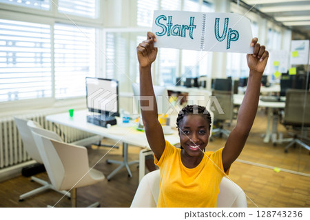 Mid adult African American woman sitting in open-plan office, holding notebook with Start Up sign Mid adult African American woman sitting in open-plan office, holding notebook with Start Up sign 128743236