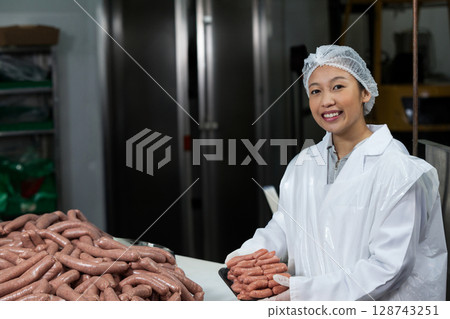 Asian woman handling tray of raw sausages on table in meat plant, wearing hairnet, copy space 128743251