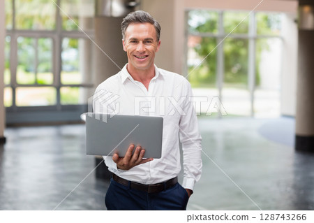 Mature adult man holding laptop in modern office lobby, showing polished floor and greenery 128743266