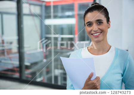Woman standing in modern office interior holding white documents and smiling, copy space 128743273