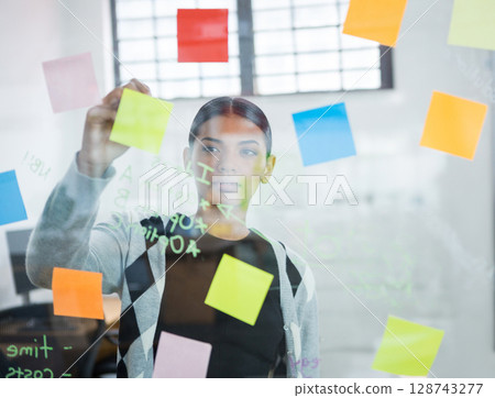 Woman arranging colorful sticky notes on glass board in open-plan office, with monitors and chairs Woman arranging colorful sticky notes on glass board in open-plan office, with monitors and chairs 128743277