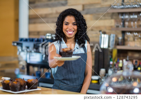 Young adult woman barista serving chocolate muffin on counter in cafe, featuring espresso machine 128743295