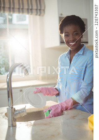 African American woman washing dishes at kitchen sink using green sponge in pink gloves, copy space 128743307