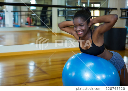 African American woman leaning on bright blue exercise ball in fitness studio, smiling, copy space African American woman leaning on bright blue exercise ball in fitness studio, smiling, copy space 128743320