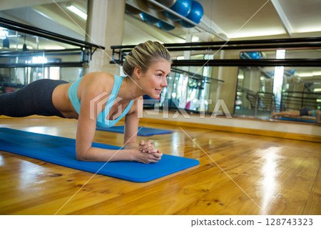 young adult woman holding forearm plank on blue mat in fitness studio with medicine ball rack 128743323