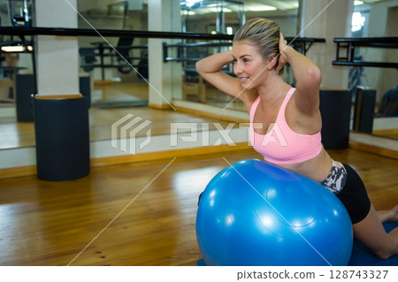 Young adult woman kneeling behind blue exercise ball in fitness studio, holding hair up, copy space Young adult woman kneeling behind blue exercise ball in fitness studio, holding hair up, copy space 128743327