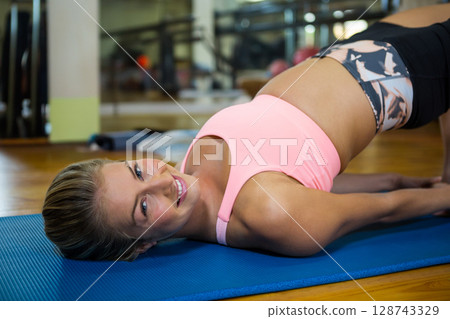 Woman lifting hips in bridge pose on blue yoga mat in studio with mirrored wall reflection 128743329