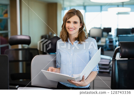 Young adult woman smiling and holding white binder in open-plan office, with cubicle partitions 128743336