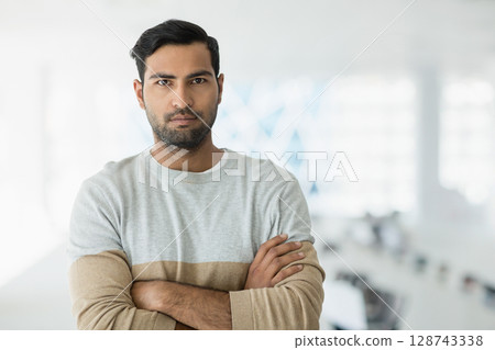 Young adult Asian man standing with arms crossed in modern open-plan office, wearing gray sweater 128743338