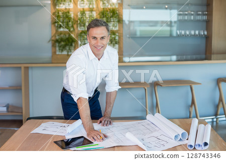 Man reviewing blueprints on wood table in modern design studio, using tablet and pencils 128743346