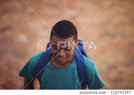 African American teenage boy walking outdoors on sunlit tan walkway, with blue backpack, smiling African American teenage boy walking outdoors on sunlit tan walkway, with blue backpack, smiling 128743347