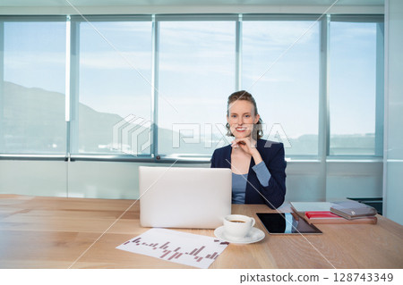 Woman sitting at meeting table overlooking waterfront, using laptop and tablet with coffee cup Woman sitting at meeting table overlooking waterfront, using laptop and tablet with coffee cup 128743349