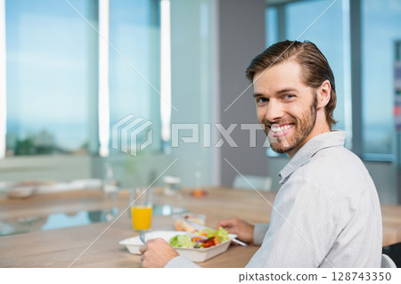 30s male eating salad from takeout box and drinking orange juice at office cafeteria, copy space 30s male eating salad from takeout box and drinking orange juice at office cafeteria, copy space 128743350
