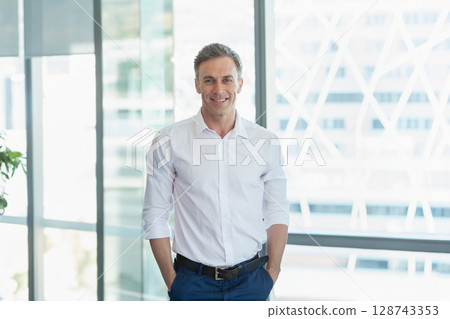 Man standing in modern office by glass windows, with potted plant and city building cross-brace Man standing in modern office by glass windows, with potted plant and city building cross-brace 128743353
