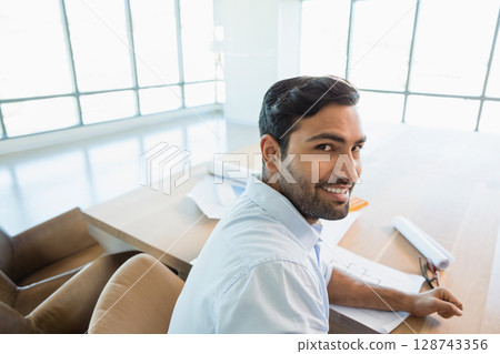 Young adult Asian man sitting at wooden desk in modern office, reviewing blueprints and smiling 128743356