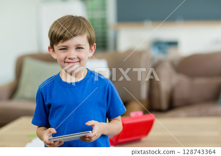 Chinese preschooler standing in living room, holding tablet near red toy container and sofa pillows 128743364