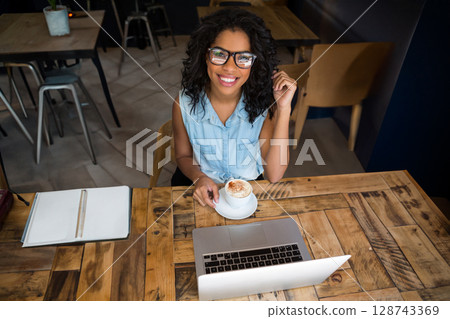 Young woman sitting at rustic café table, wearing glasses and holding coffee cup while using laptop 128743369