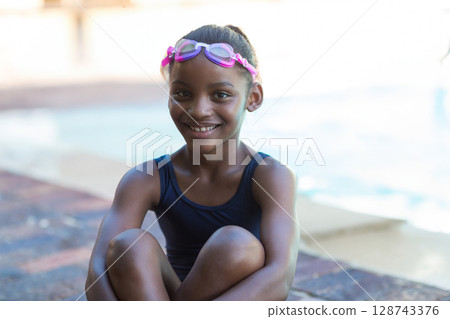 African American girl sitting cross legged at outdoor poolside, wearing swim goggles and swimsuit African American girl sitting cross legged at outdoor poolside, wearing swim goggles and swimsuit 128743376