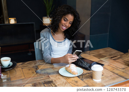 Woman sitting at wooden table in cozy café, using smartphone and tablet with latte and croissant Woman sitting at wooden table in cozy café, using smartphone and tablet with latte and croissant 128743378