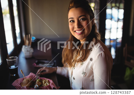 Young woman sitting at counter by window in café, eating fries from checkered basket, sipping drink 128743387