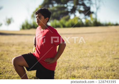 Boy performing lunge stretch on dry grassy field outdoors, with yellow wrist wrap and red T?shirt 128743395