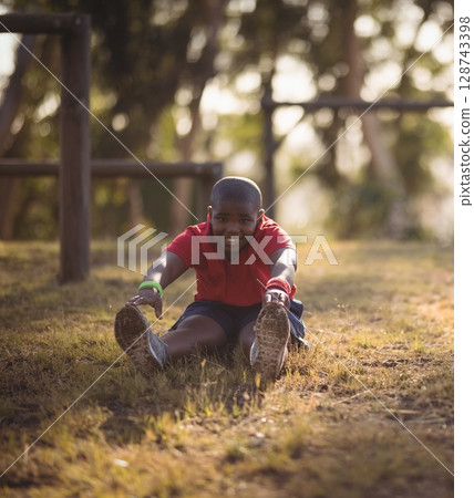 African American boy stretching forward on dry grass in park by wooden bars, wearing wristbands 128743398