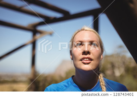 Woman standing under wooden pergola beams outdoors, looking upward toward clear blue sky 128743401