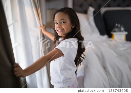Girl pulling beige curtains and smiling in bright bedroom, with white bed linens and morning light Girl pulling beige curtains and smiling in bright bedroom, with white bed linens and morning light 128743408