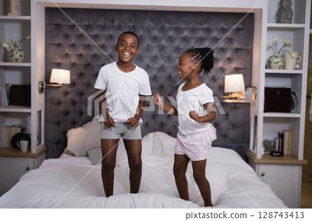 African American child siblings jumping on bed in bedroom, with headboard pillows lamps shelves 128743413