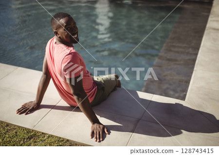 African American man sitting on pool edge at tiled deck beside grass patch, leaning and smiling 128743415