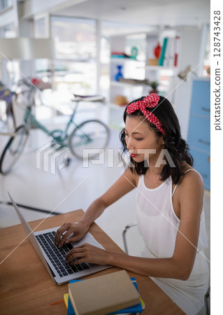 Asian woman typing on laptop at home office table, with notebooks, mint green bicycle, copy space 128743428