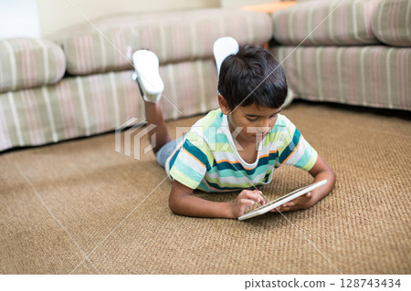 School-aged bi-racial boy lying on brown carpet in living room, tapping white tablet School-aged bi-racial boy lying on brown carpet in living room, tapping white tablet 128743434