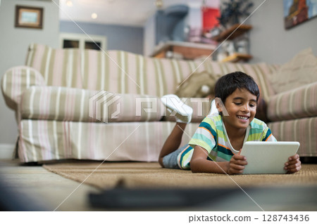 6-year-old boy lying on woven rug in living room, holding white tablet and smiling 128743436