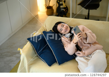 Cheerful teenage girl lying on a couch, relaxing at home and using smartphone 128744297