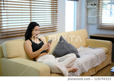 Happy teenage girl relaxing on a sofa, laughing and browsing her smartphone 128744325
