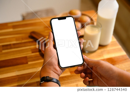 Hands holding mobile phone with empty display over wooden kitchen counter and breakfast 128744340